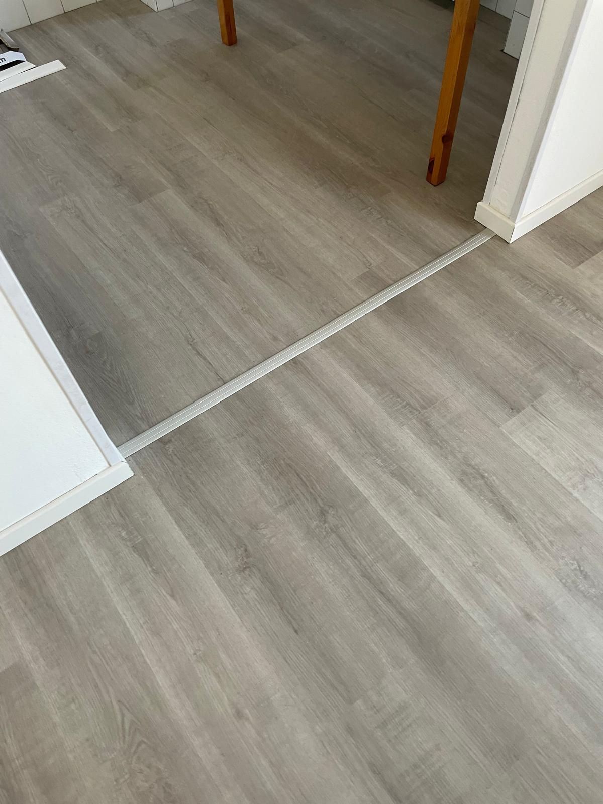 Pavimento in legno chiaro con una soglia metallica tra due stanze e un bordo bianco sulla porta.