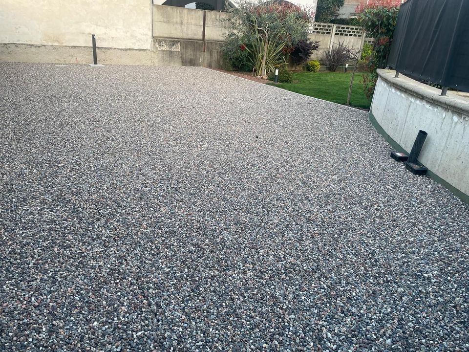 Cortile posteriore ricoperto di ghiaia adiacente a una casa, con una piccola aiuola e una recinzione sullo sfondo.