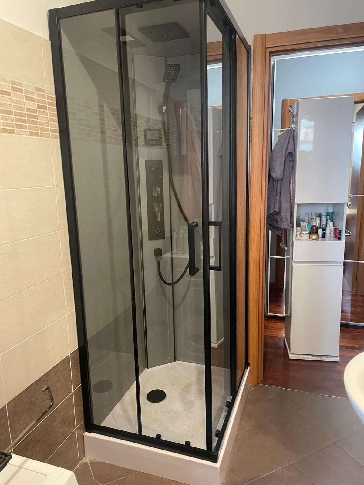 Box doccia compatto con struttura nera e porta in vetro trasparente, installato in un bagno piastrellato.