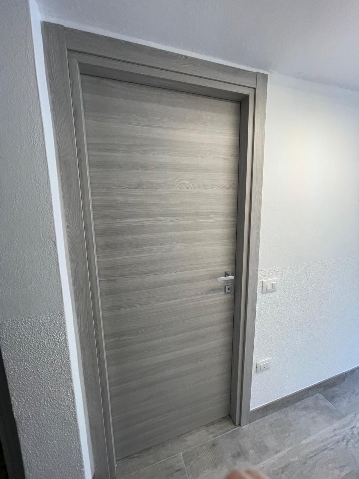 Porta interna chiusa in legno chiaro in un corridoio bianco con pavimento in piastrelle grigie.