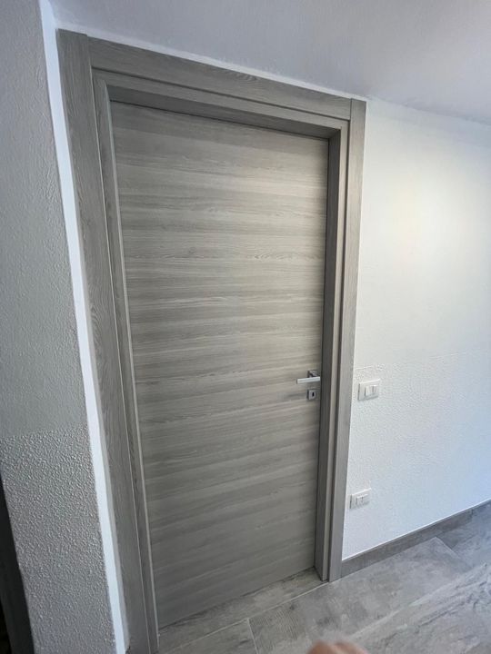 Porta interna chiusa in legno chiaro in un corridoio bianco con pavimento in piastrelle grigie.