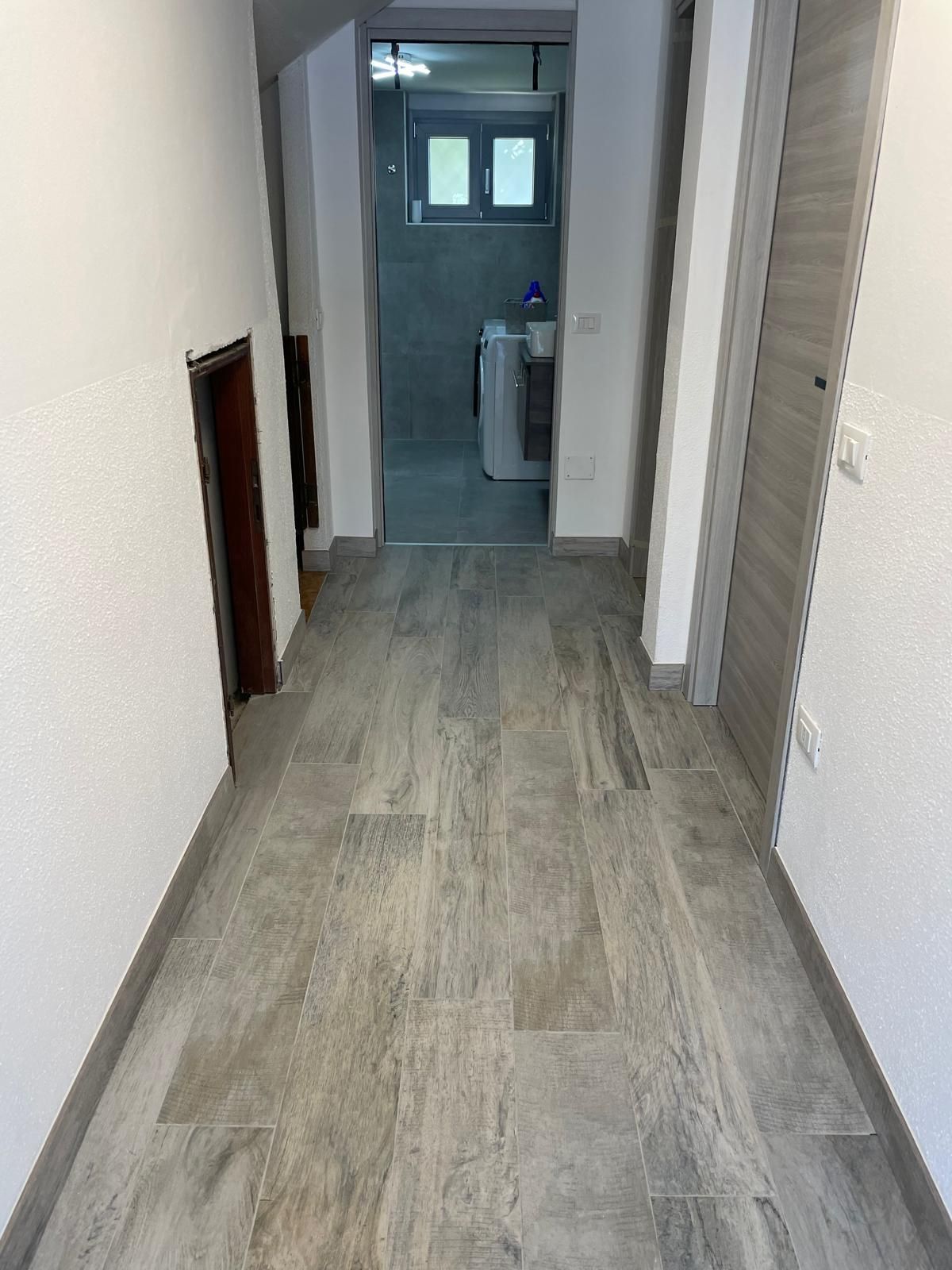 Stretto corridoio con pavimento in legno grigio che conduce a un piccolo bagno in fondo.