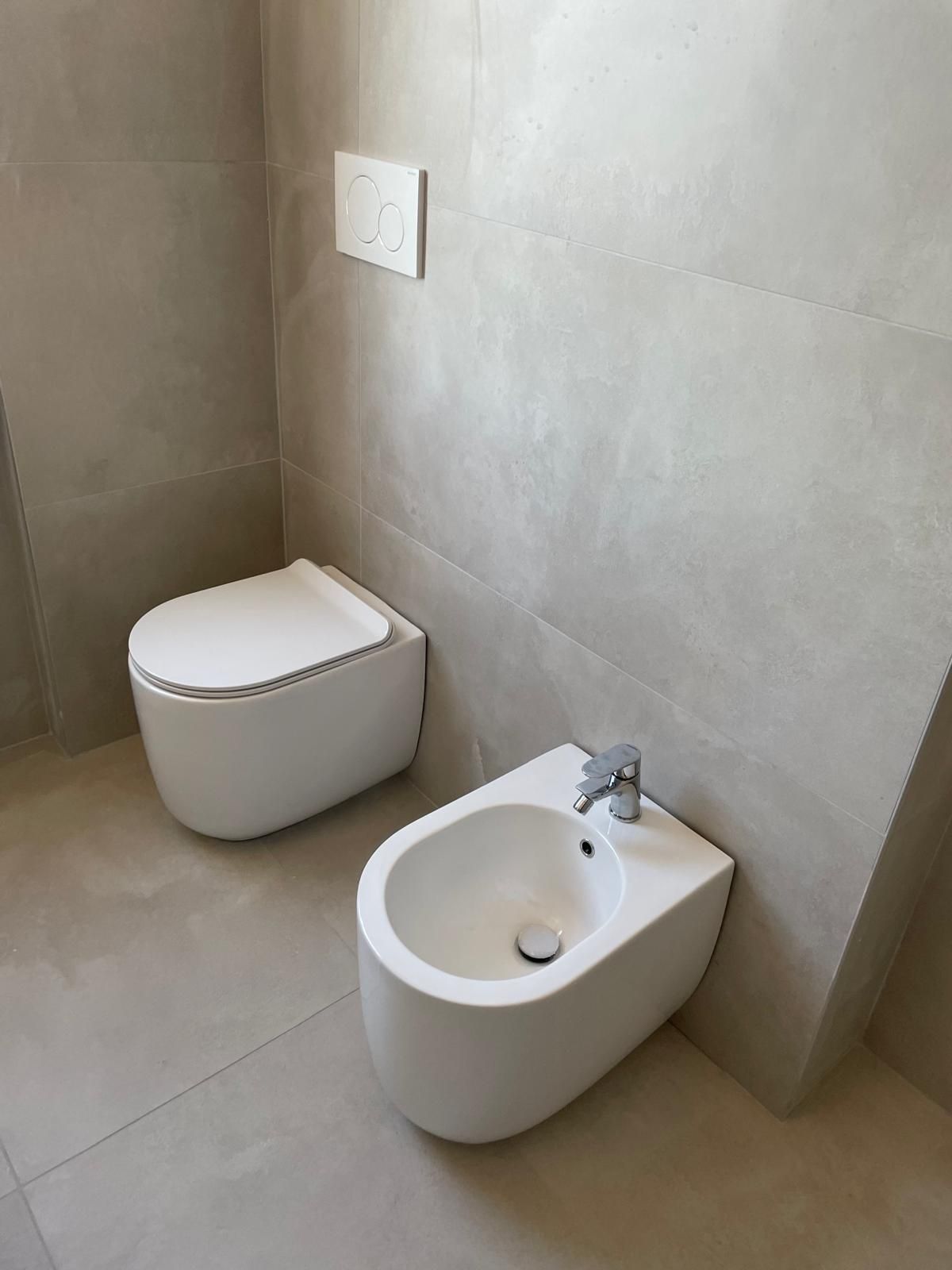 Bagno moderno con WC sospeso e bidet in una stanza piastrellata di grigio.