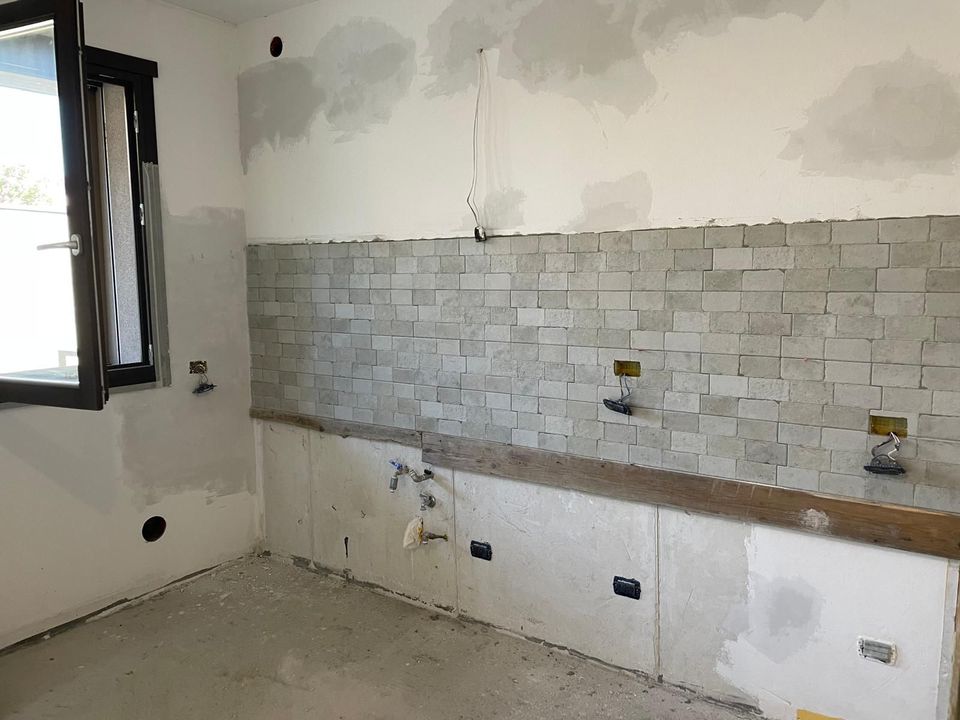 Cucina in fase di ristrutturazione con paraschizzi piastrellato, pareti bianche non rifinite e una finestra sulla sinistra.