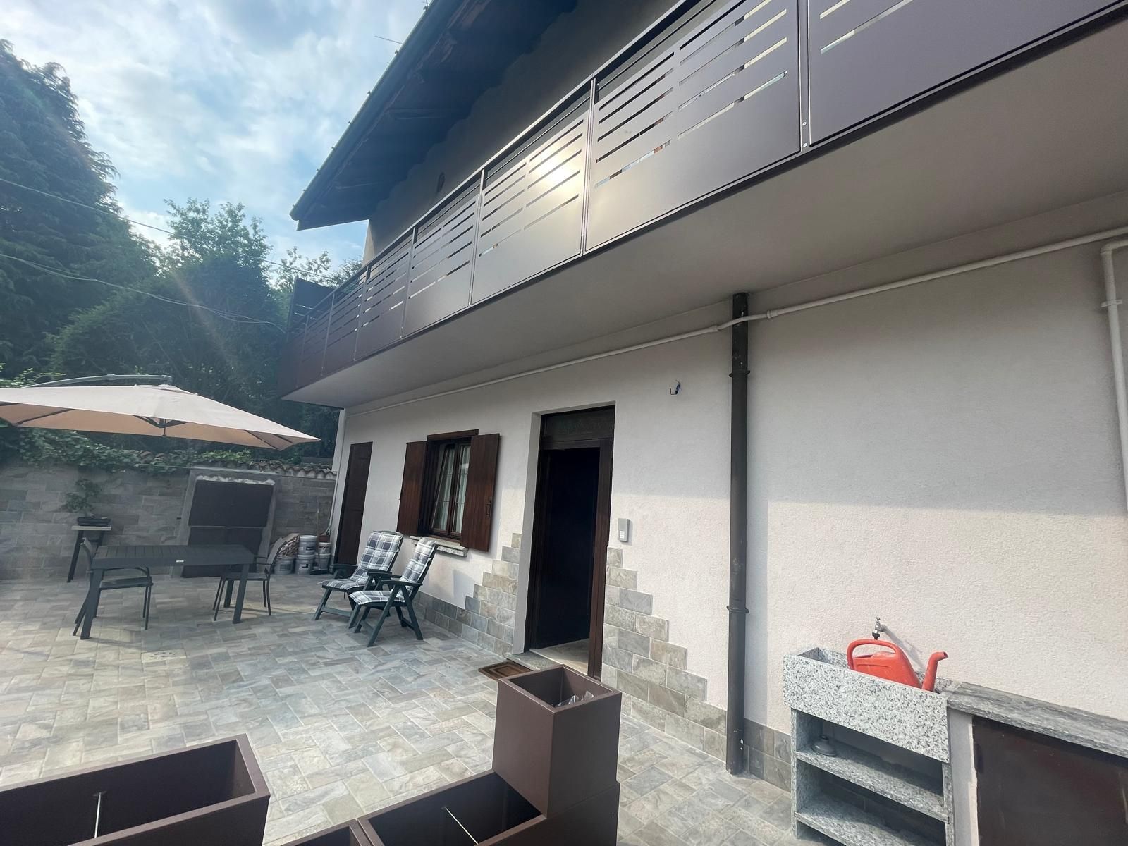 Patio esterno adiacente alla casa, con sedie, tavoli e una porta aperta sotto un balcone coperto.
