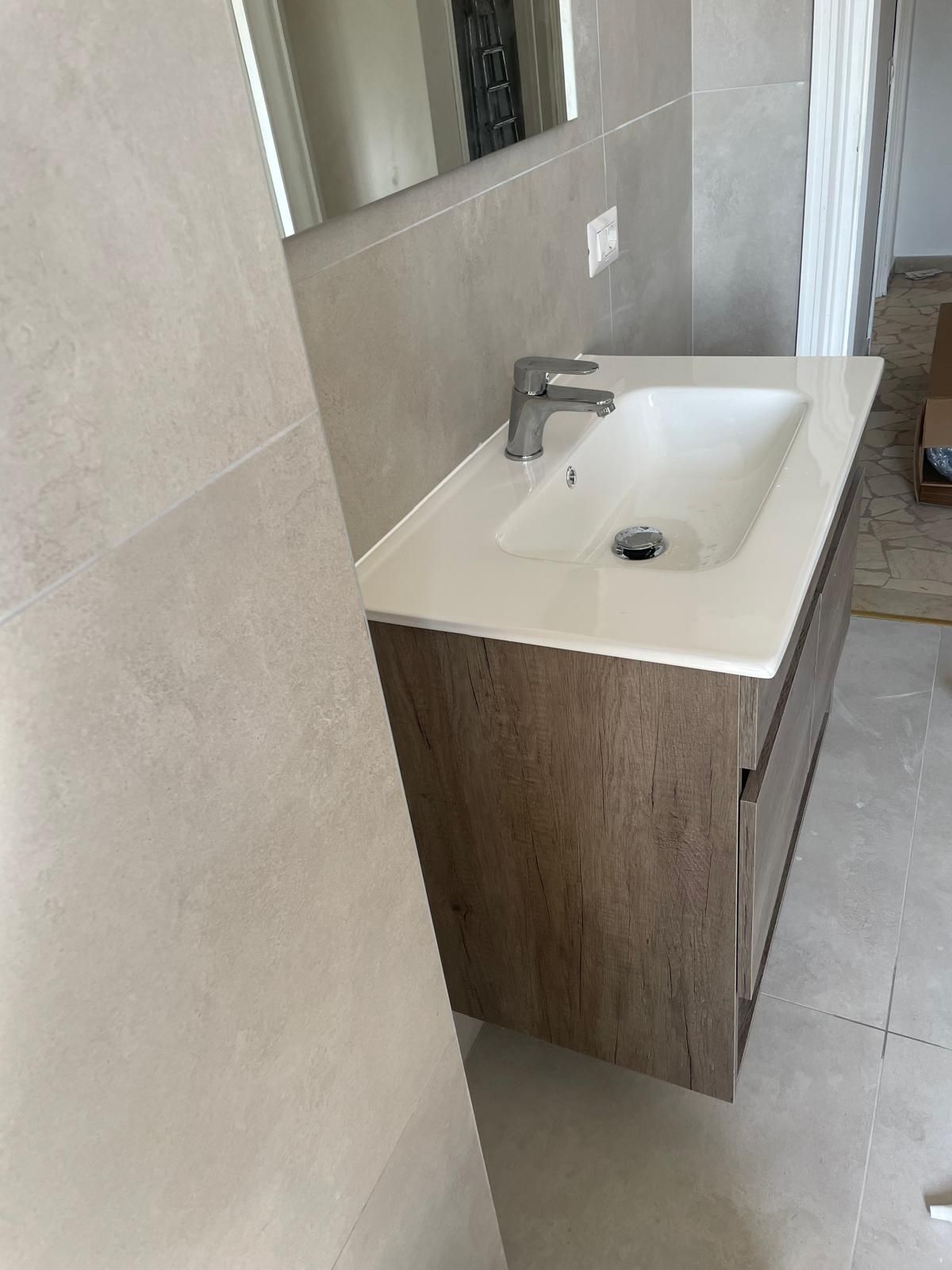 Mobiletto da bagno di piccole dimensioni con lavabo e specchio bianchi, su sfondo di piastrelle beige e mobiletto in legno.