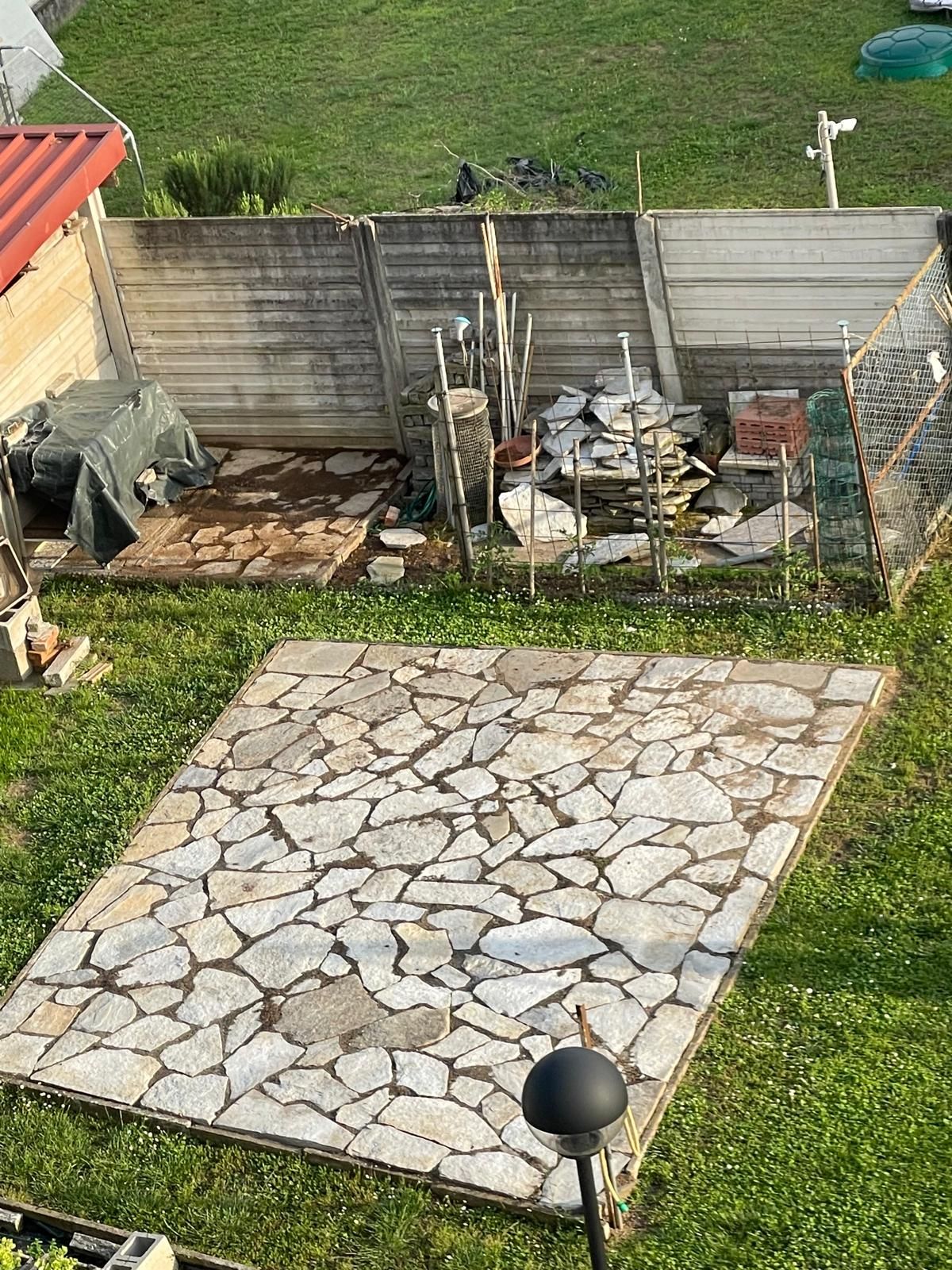Patio sul retro con una piazza pavimentata in pietra e attrezzi da giardino lungo la recinzione.