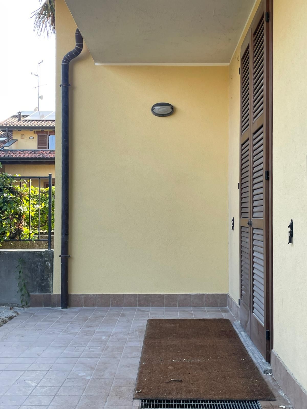 Veranda coperta con parete beige, pavimento piastrellato, applique e porte a persiana in legno accanto a uno zerbino.