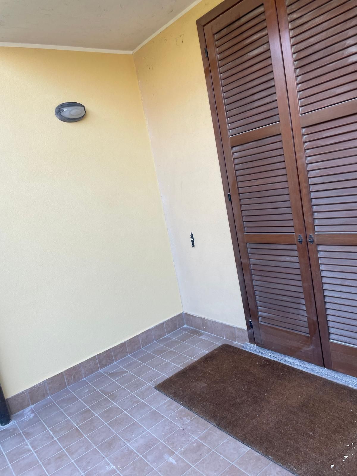 Veranda con parete beige, porta con persiane marroni, applique e zerbino su pavimento piastrellato.