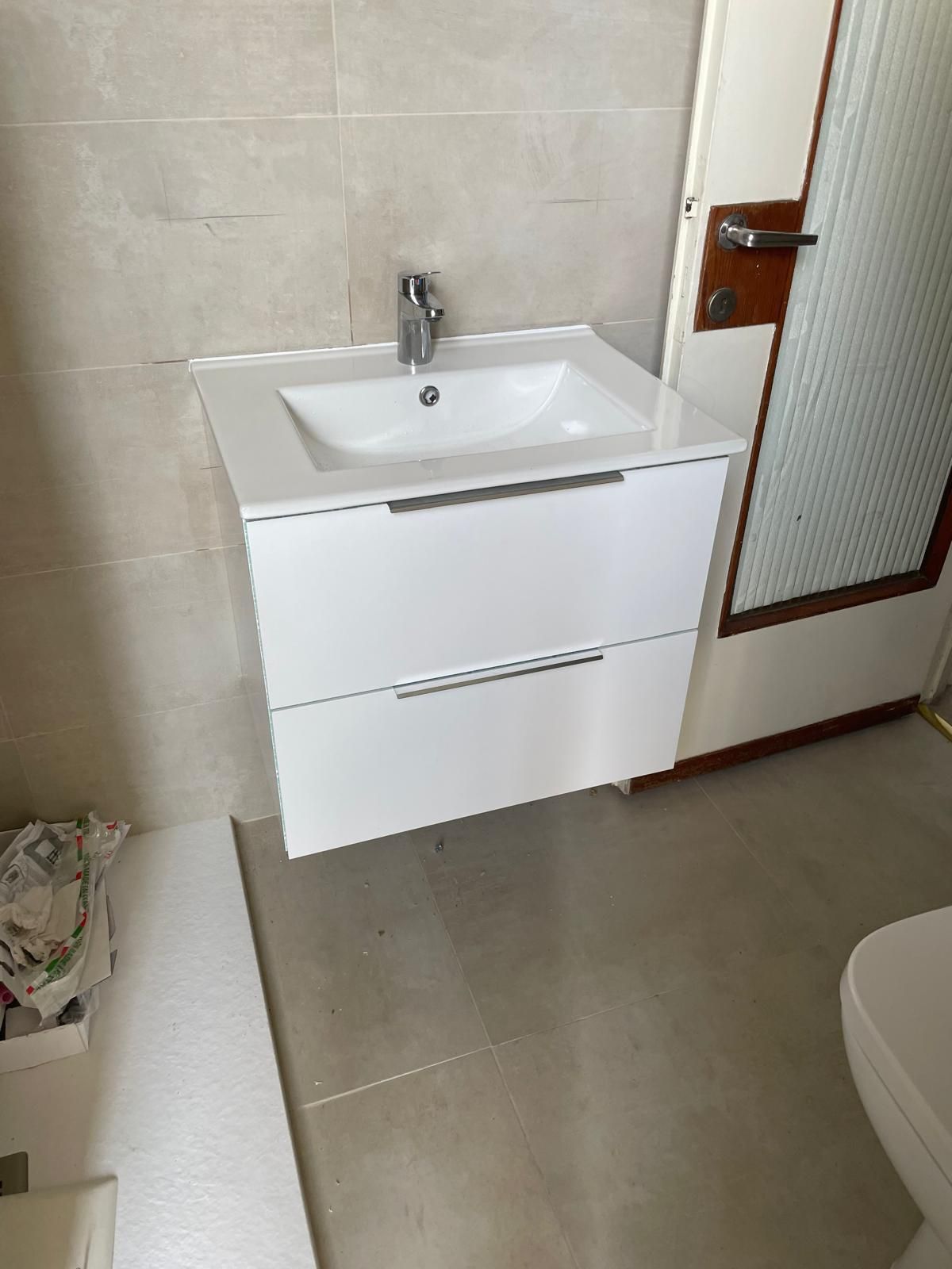 Piccolo mobiletto da bagno con lavabo bianco accanto a una porta, pareti e pavimento piastrellati.