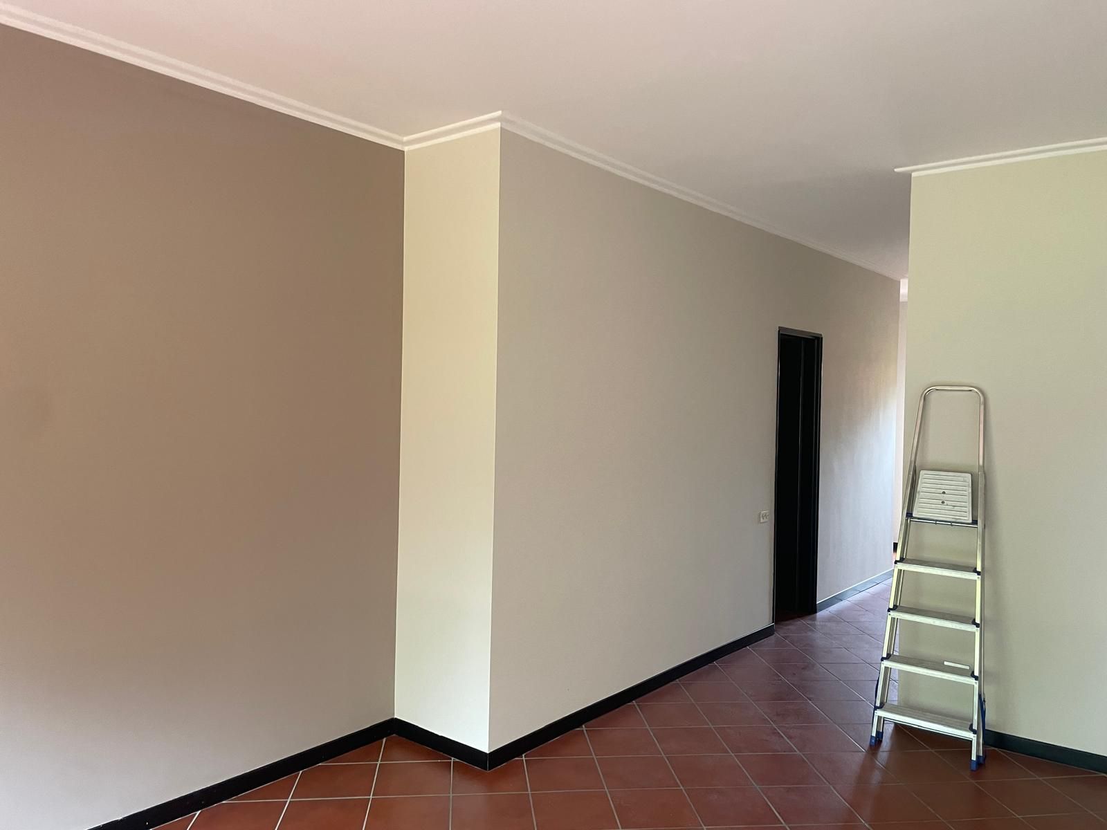 Stanza vuota con pareti beige, finiture nere e una scaletta vicino a una porta scura.