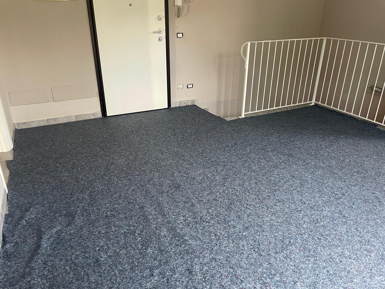 Stanza vuota con moquette blu, pareti bianche e una ringhiera sul lato destro.