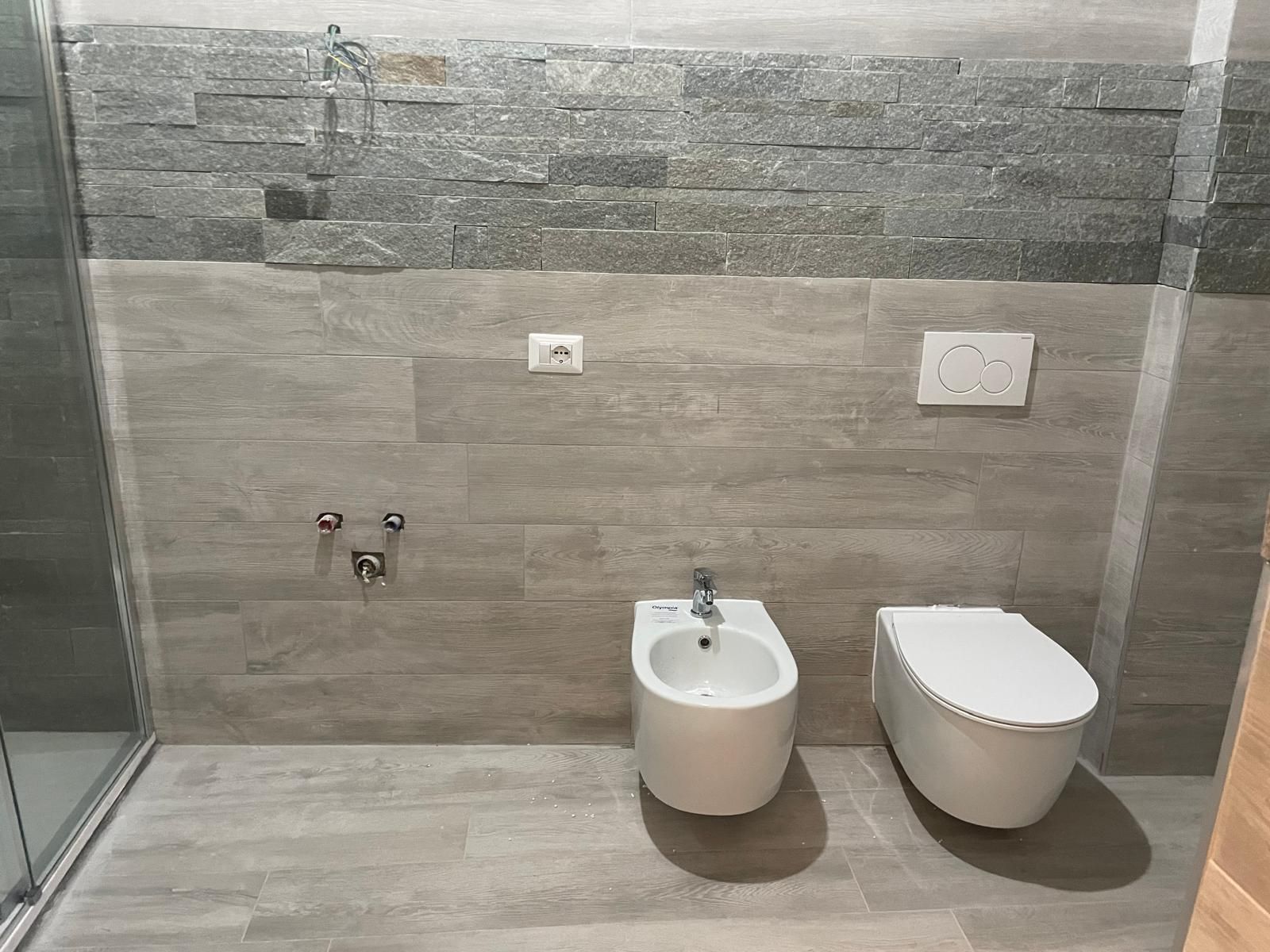 Bagno moderno piastrellato con lavabo a parete, WC e bidet in tonalità di grigio.