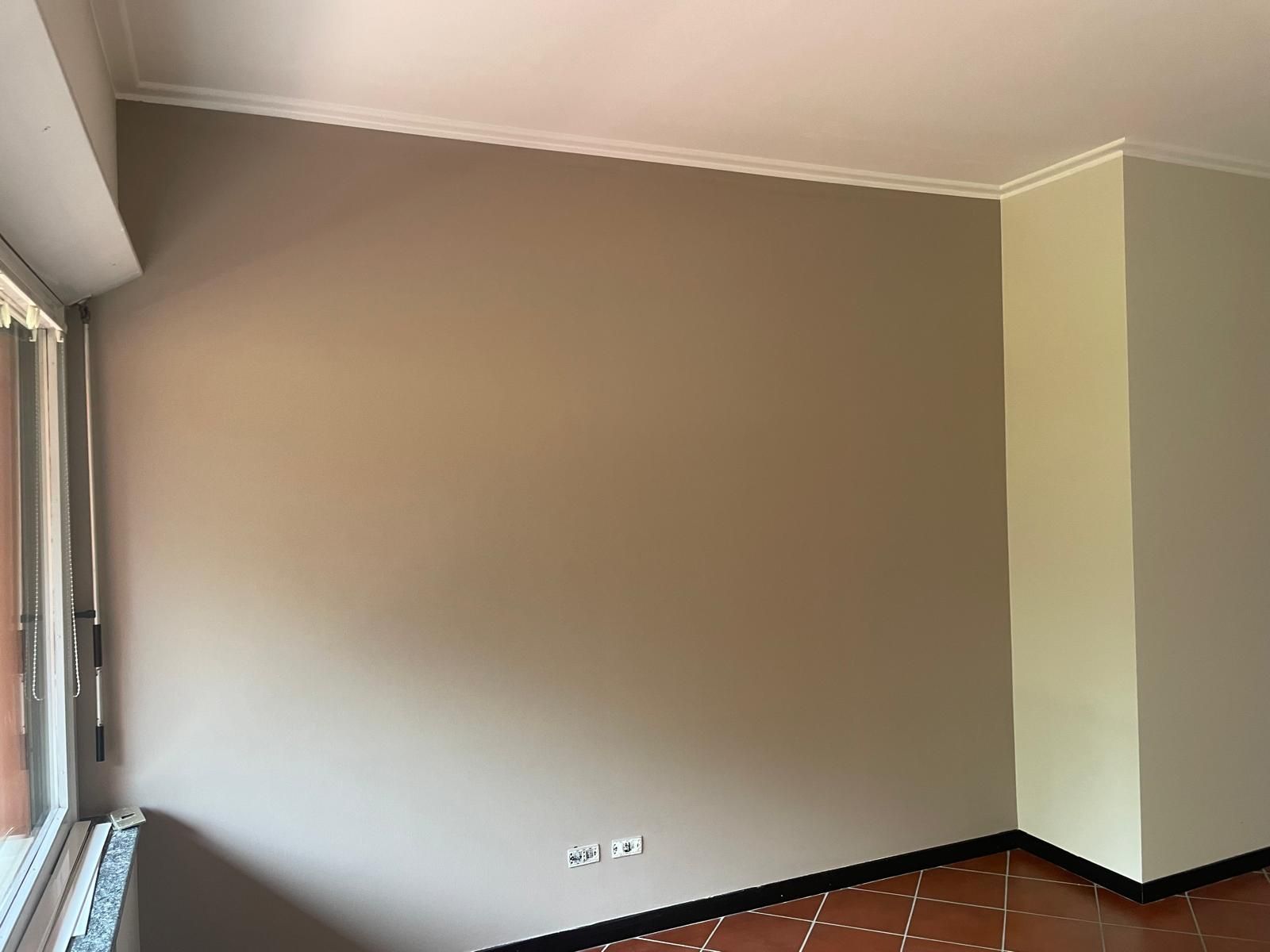 Stanza vuota con pareti beige, battiscopa neri e una finestra sulla sinistra.