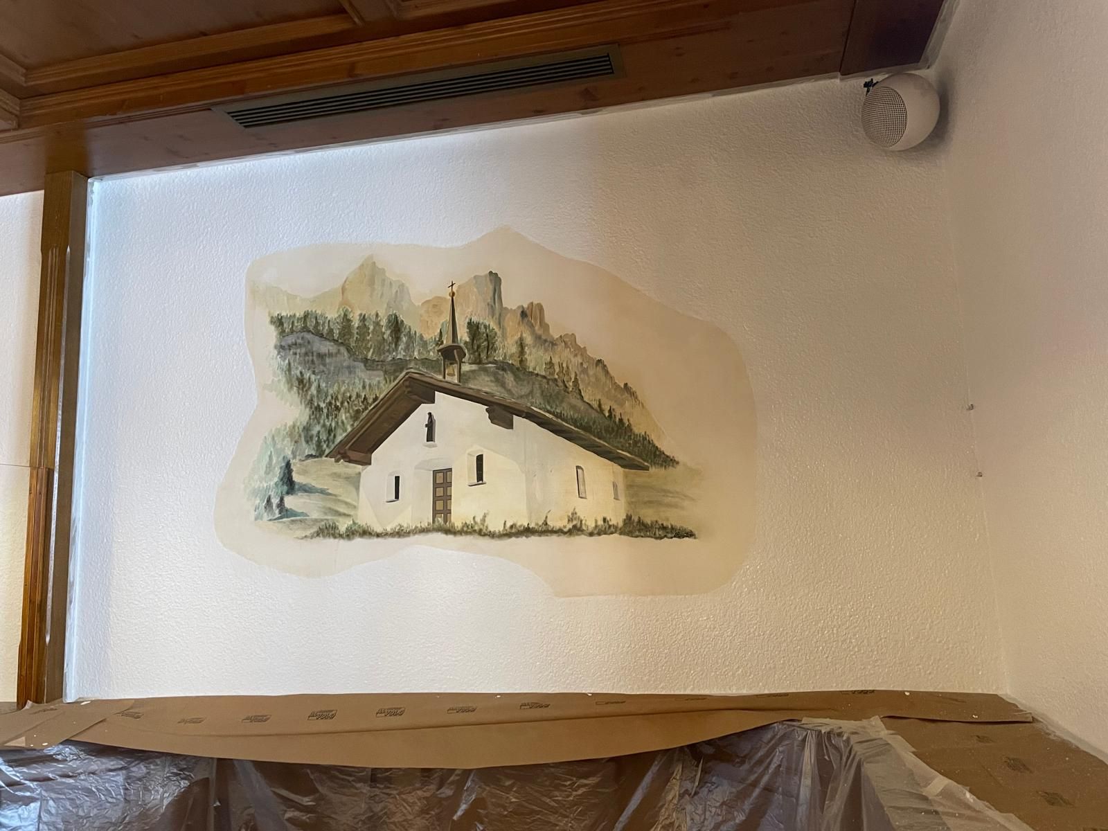 Piccolo dipinto murale raffigurante una casa bianca con tetto spiovente e alberi, appeso all'interno sopra una mensola di legno.