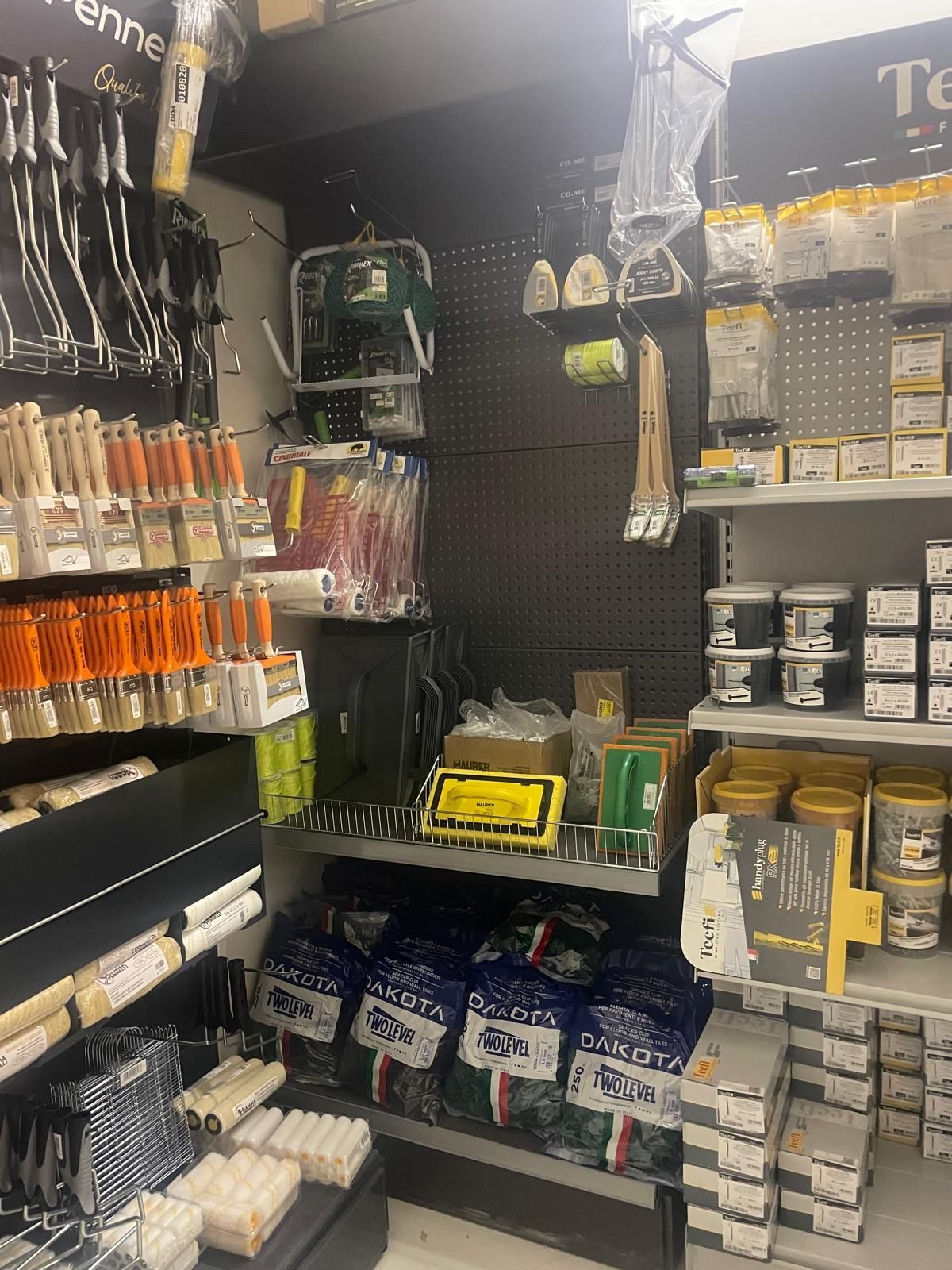 Corridoio di un negozio di ferramenta con attrezzi appesi, forniture in scatola e scaffali di rulli per pittura e elementi di fissaggio.