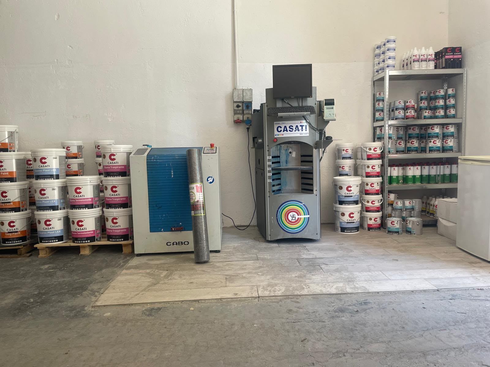 Corridoio del magazzino con secchi di vernice impilati, una macchina per miscelare e scaffalature metalliche.