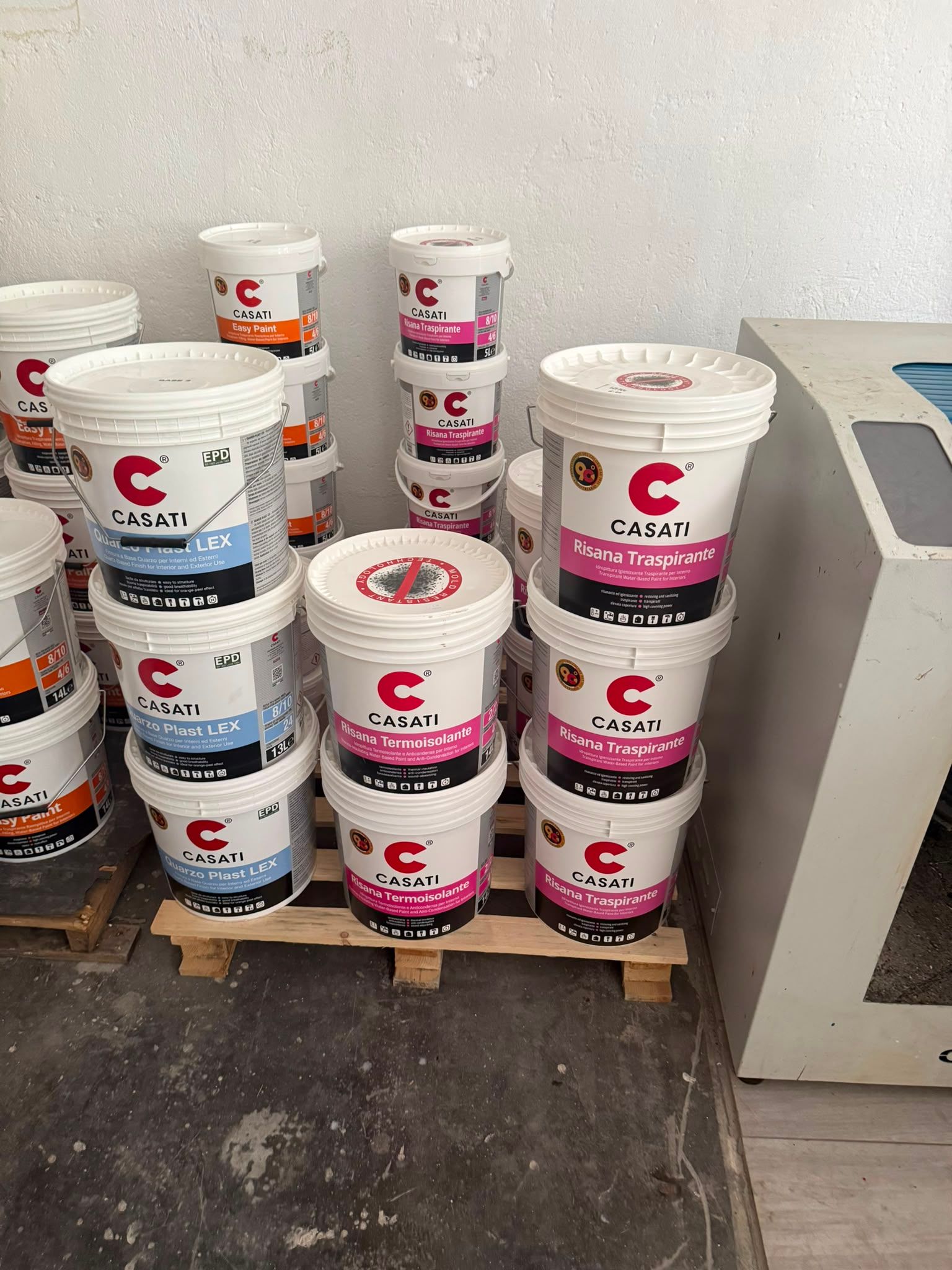 In un'officina, accanto a un macchinario, sono impilati dei secchi di vernice bianca con etichette rosa e blu su un pallet.