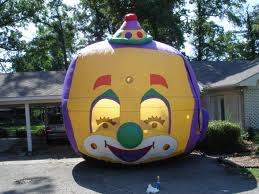 Giant inflatable clown face with large eye openings, yellow and purple with a green nose, in front of a house.