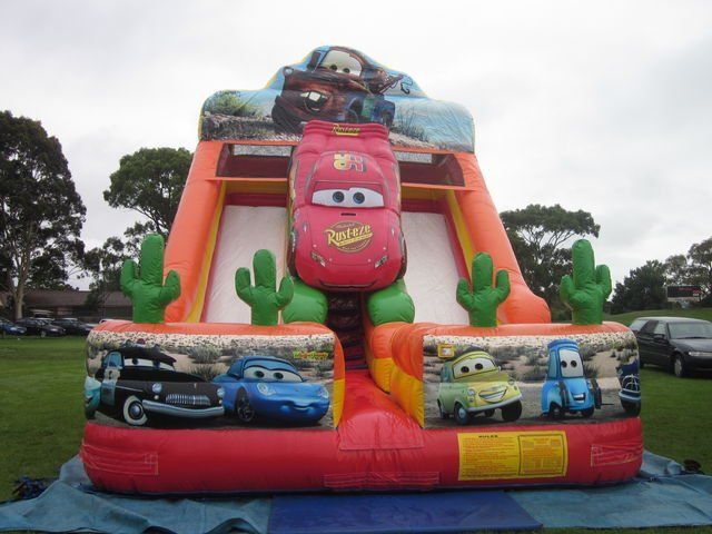 Cars-themed inflatable slide with Lightning McQueen, cacti, and Cars characters in a park setting.