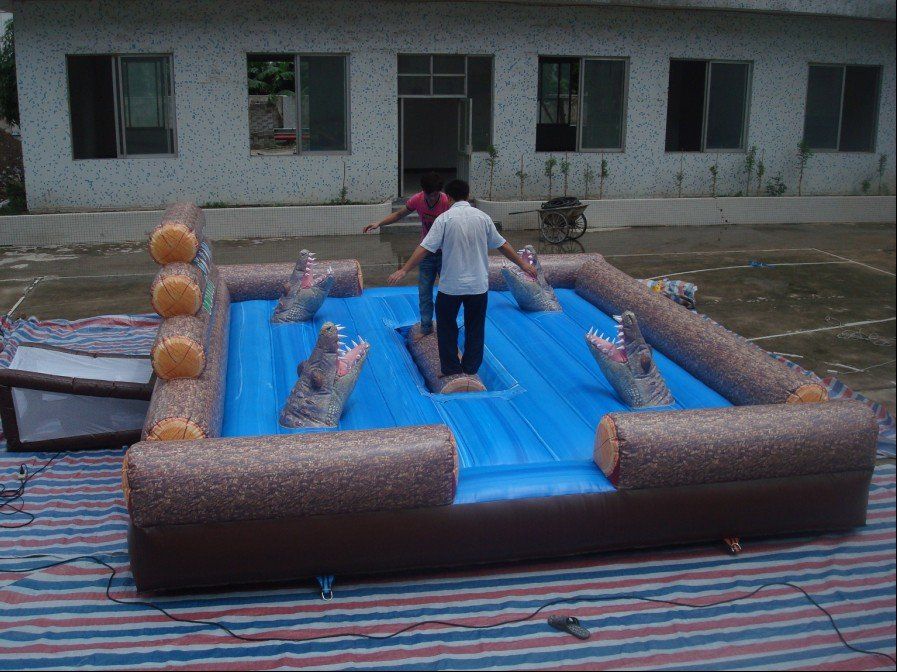A person balances on an inflatable log bridge above inflatable crocodiles in a play area.