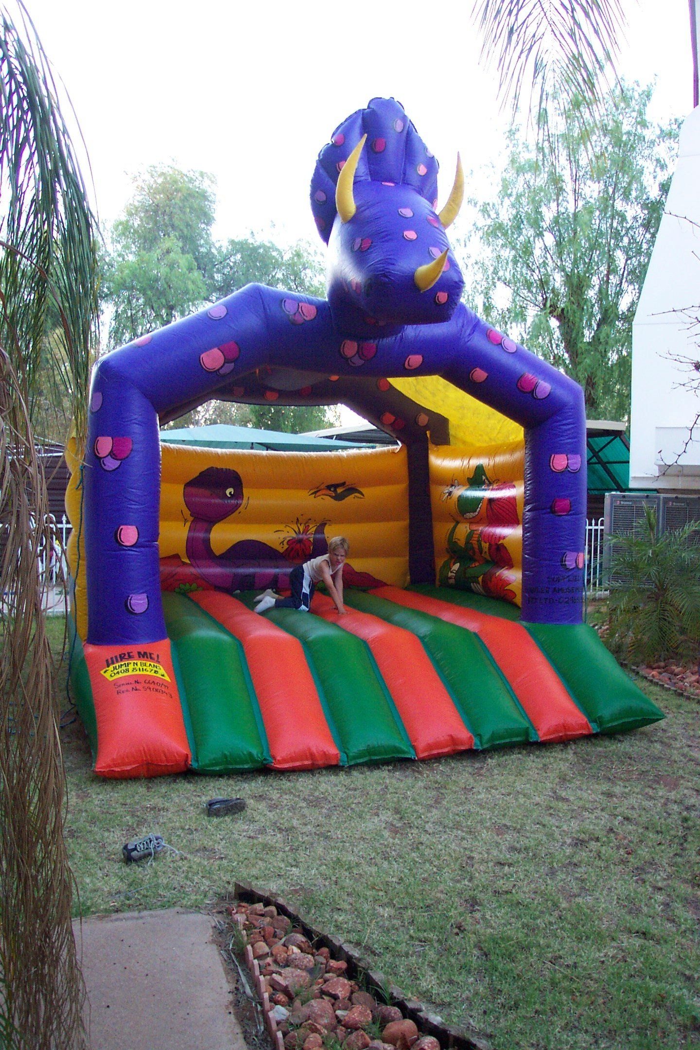 Bouncy house shaped like a purple elephant with green and red stripes on the jumping platform, set outdoors in a yard.