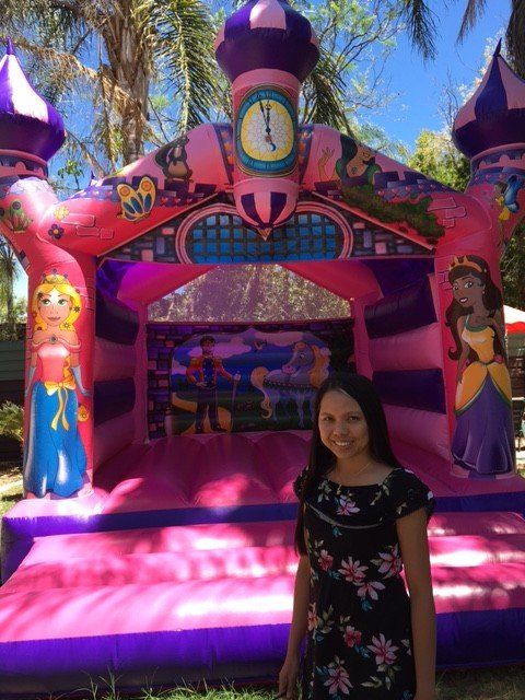 Woman standing in front of a pink and purple princess-themed bounce house outdoors on a sunny day.