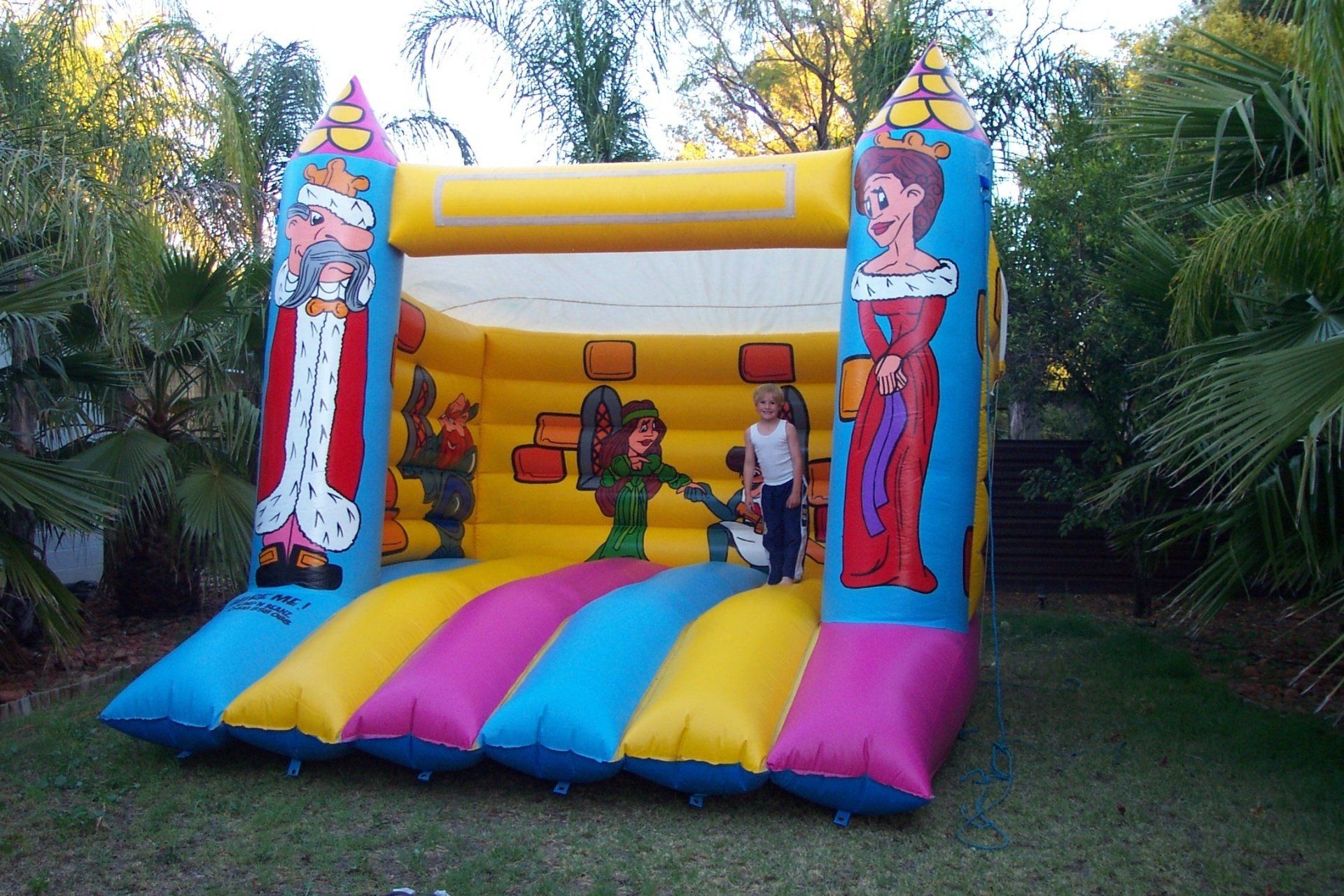 A colorful inflatable bounce house with a princess and knight theme set on grass, with two children inside.