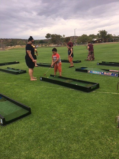 A boy plays mini golf on a green field, watched by adults and other children under an overcast sky.