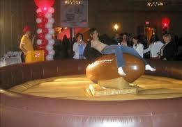Woman riding a mechanical bull at an indoor event. She is in jeans and is holding on. A crowd watches.
