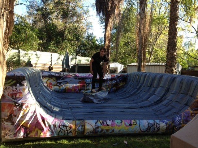 A person on a skateboard in a graffiti-covered inflatable skate park. The setting is outdoors, amidst trees and grass.