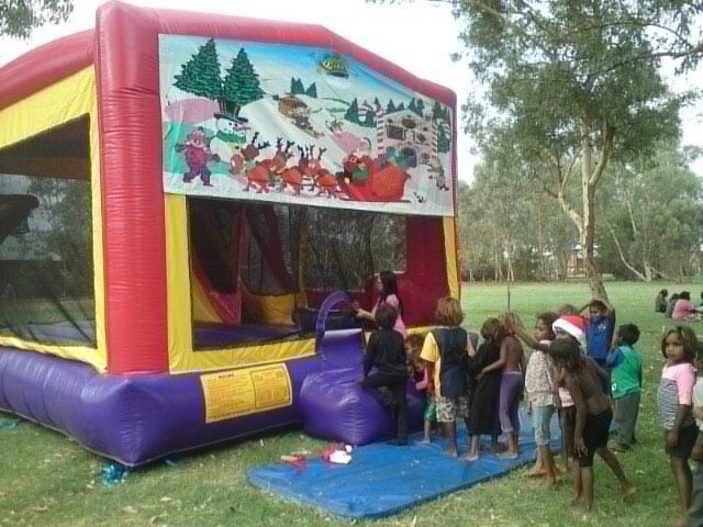 Children line up to enter a colorful Christmas-themed bouncy castle outdoors on a grassy field.