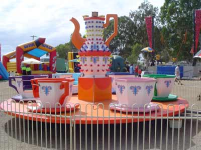 A carousel ride with teacups around a giant teapot. The ride is on a platform, surrounded by a fence, at an outdoor carnival.