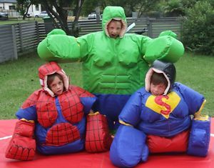 Three children in inflatable superhero costumes: Hulk, Spider-Man, and Superman, on a red mat outdoors.