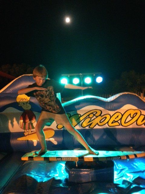 Boy balancing on a mechanical surfboard at night, with a full moon visible. He's on a WipeOut inflatable with spotlights.