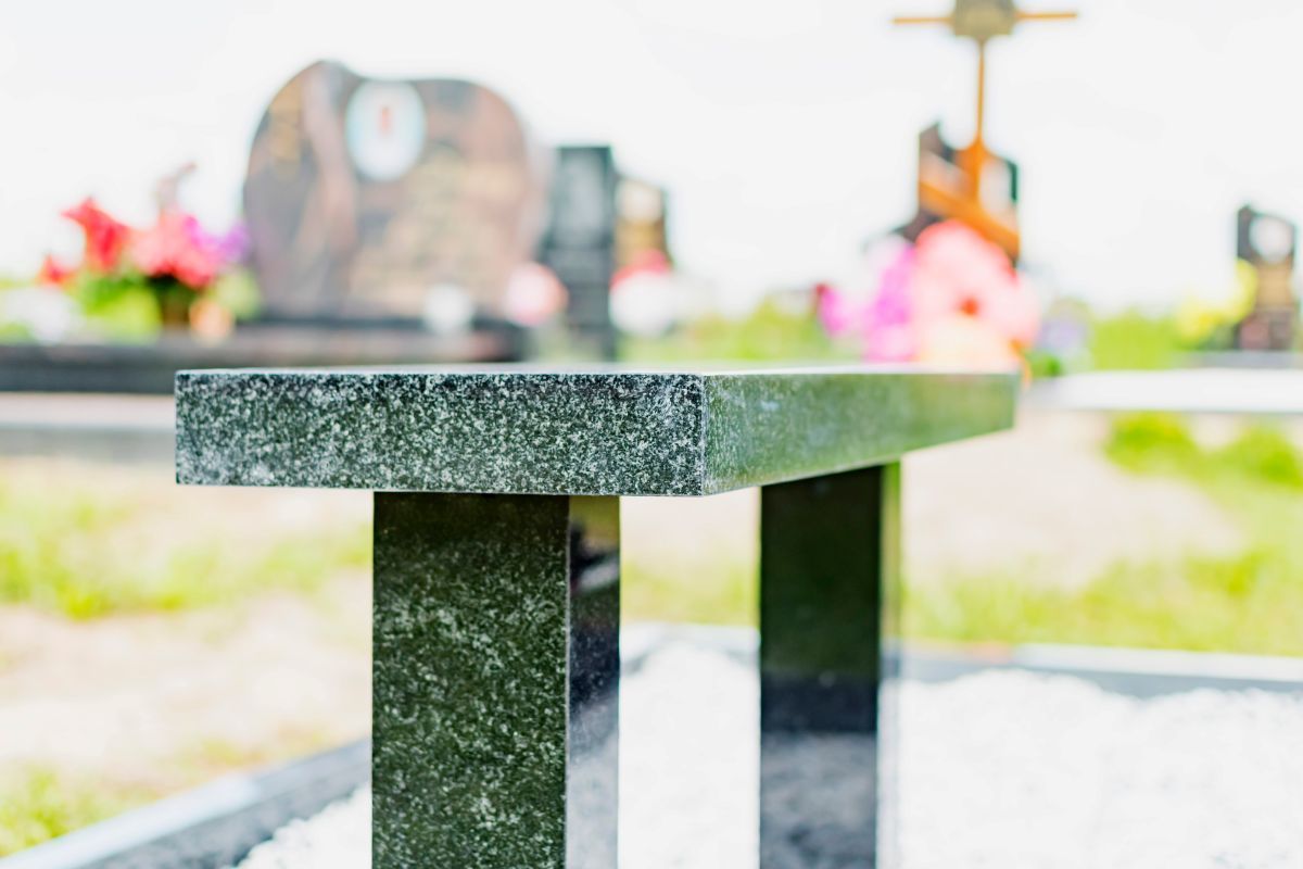 A black granite bench in a cemetery, with headstones and flowers blurred in the background.