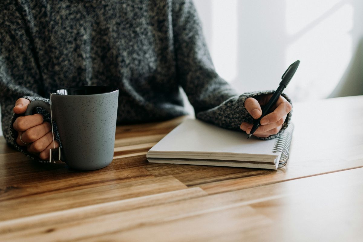 Person writing in notebook with pen at a wooden table, holding a coffee mug. Gray sweater and mug.