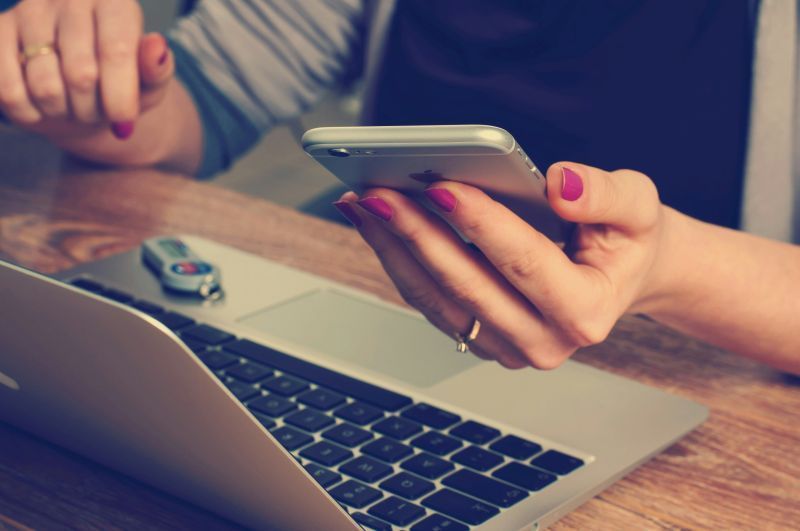 Woman using a smartphone and laptop at a wooden desk. She has pink nail polish and wears a ring.
