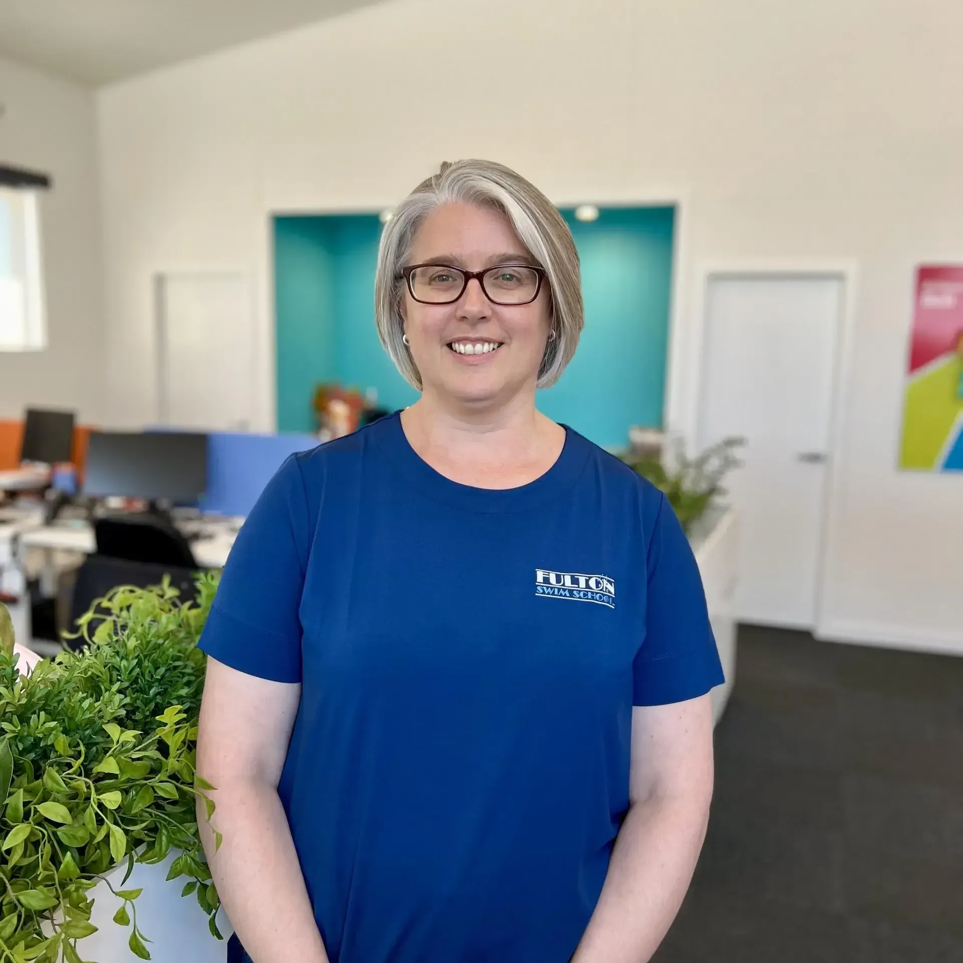 A woman wearing glasses and a blue shirt is standing in an office.