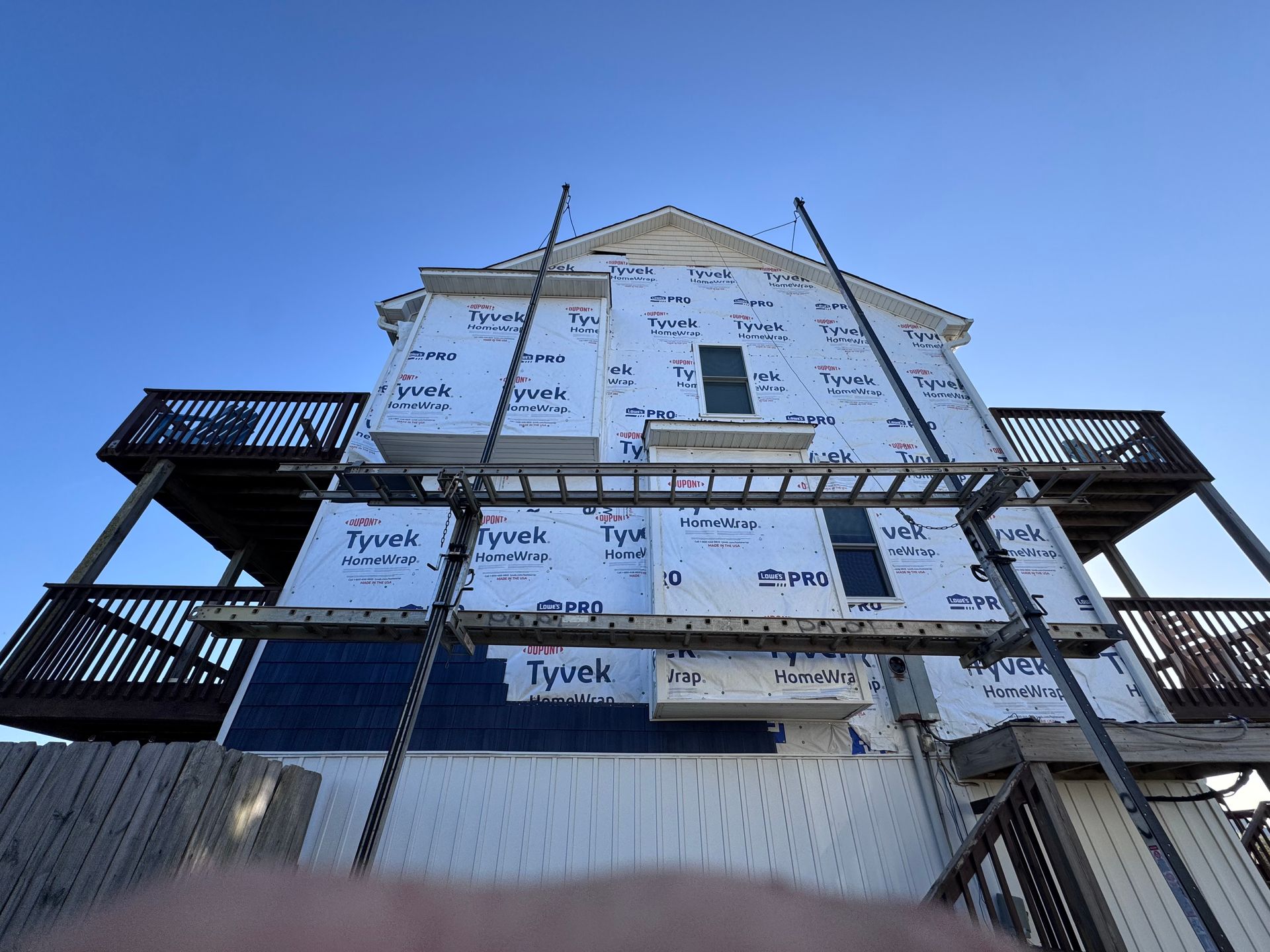 House exterior under construction, covered in Tyvek wrap, with ladders and scaffolding.