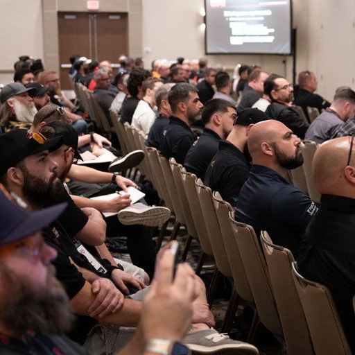 Audience in rows of chairs at a conference, looking at a screen, listening.
