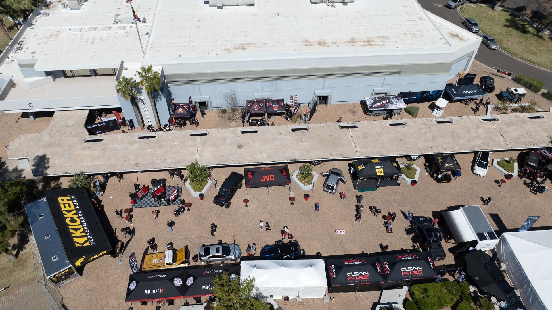 Aerial view of an event outside a large building with cars, booths, and people.