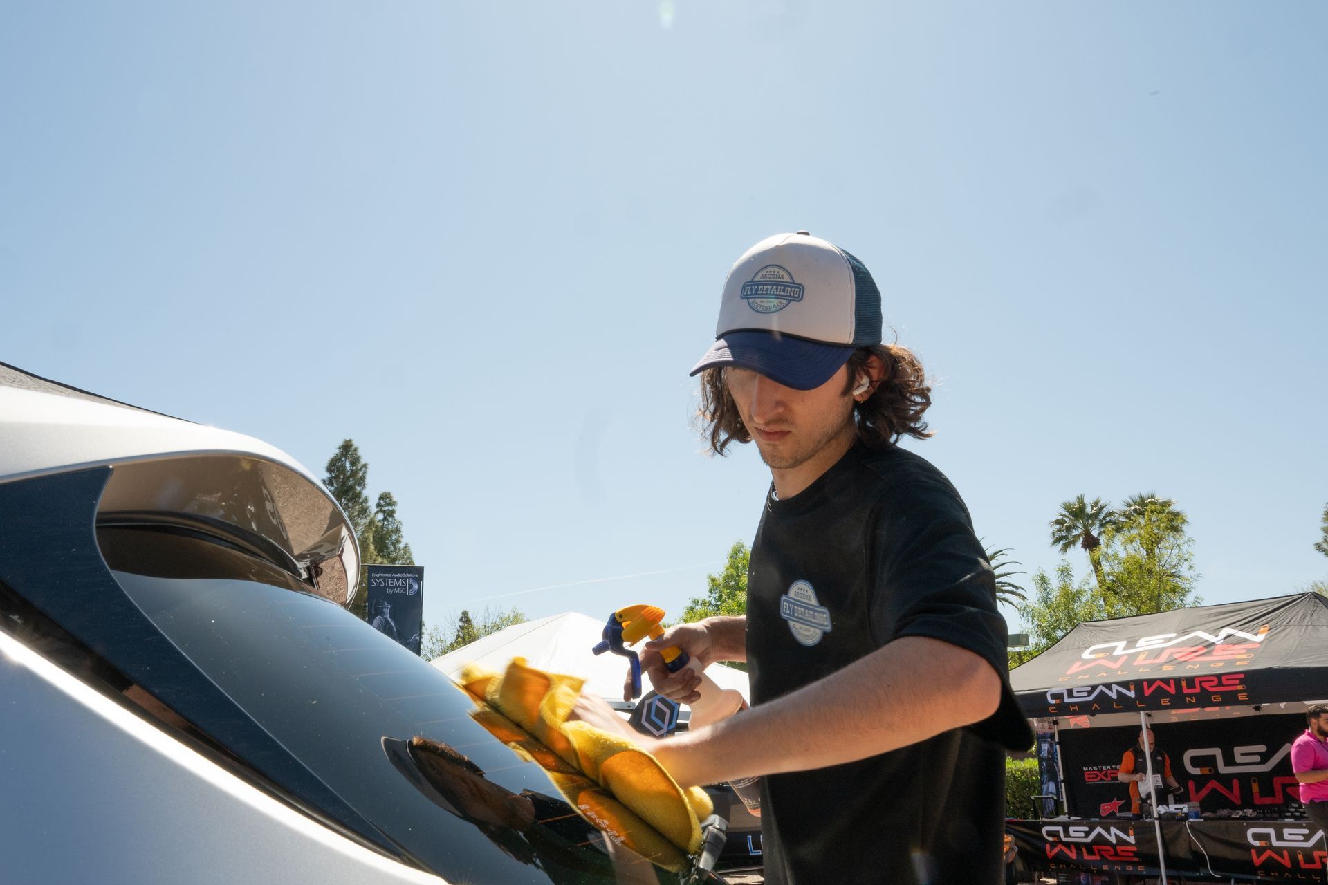 Man cleaning a car window with a spray bottle and cloth on a sunny day.