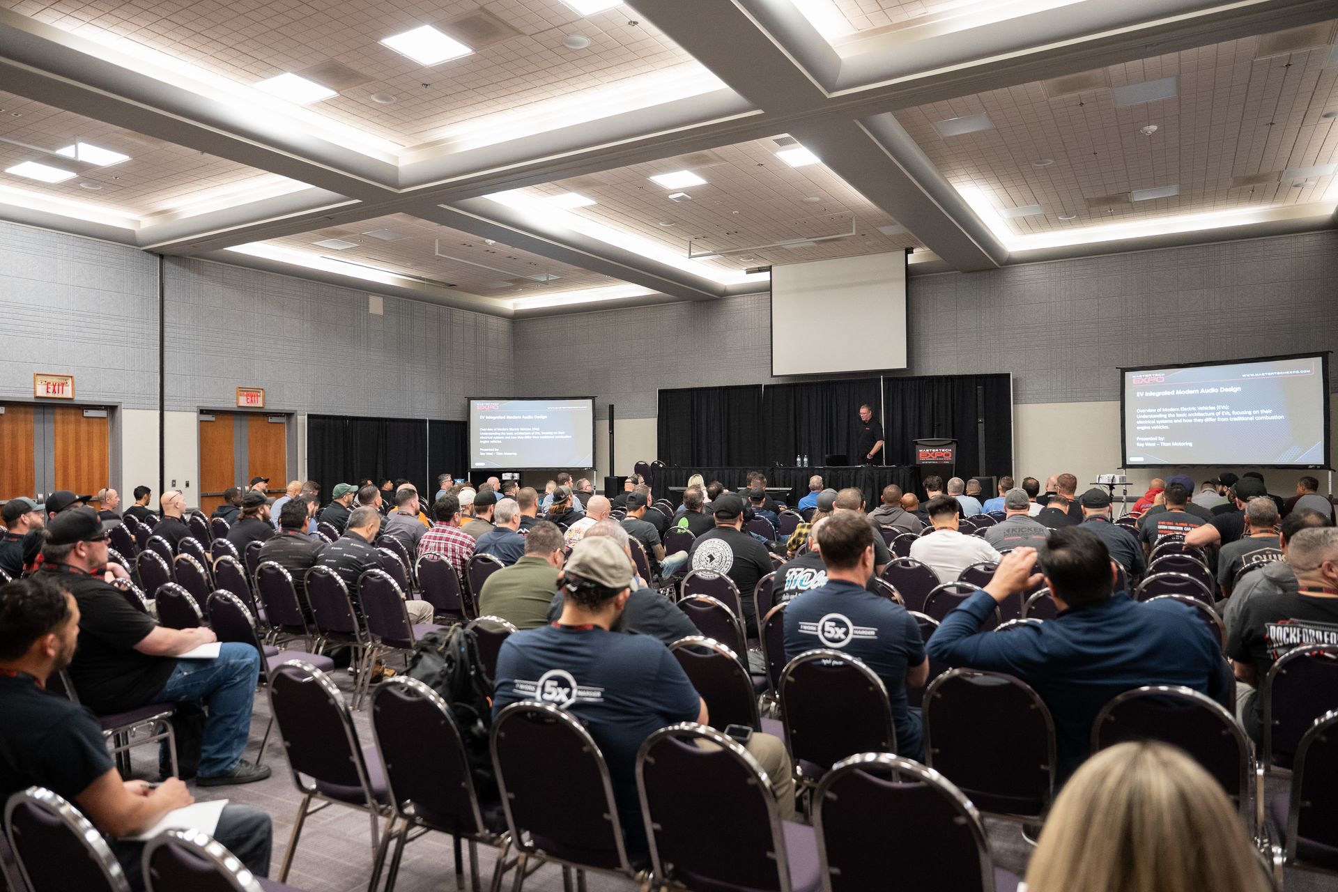 Audience at a conference presentation; speaker at podium, screens display code.