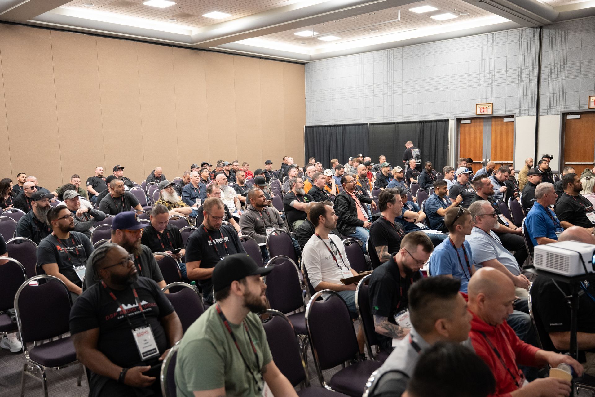 Audience in a conference room; people seated, listening to a presentation. Projector, muted tones, neutral lighting.
