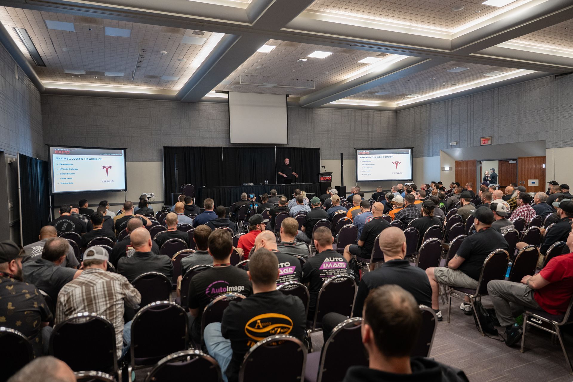 Audience at a conference presentation; large room, people in chairs facing a stage with screens.