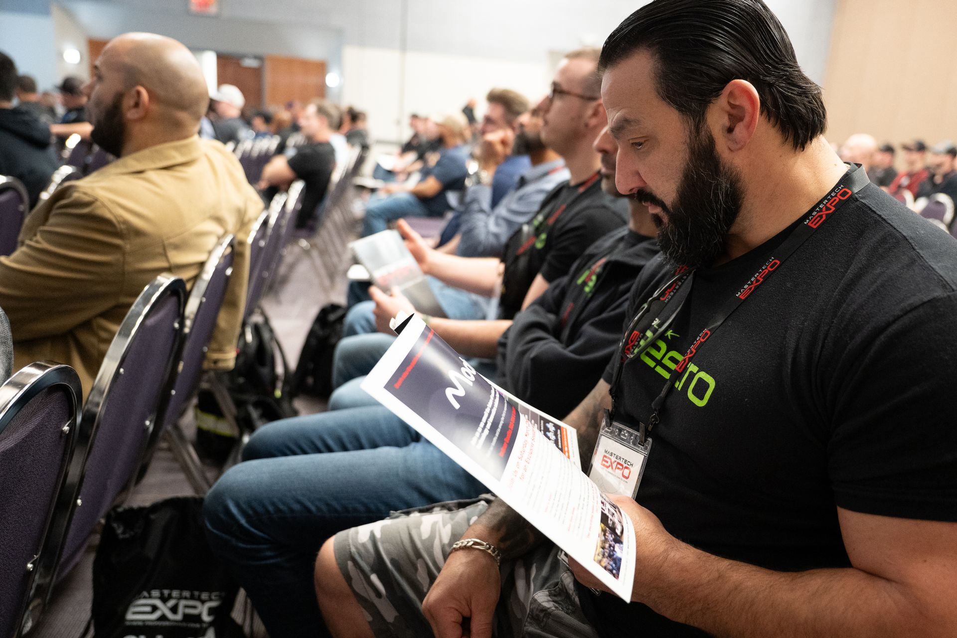 Attendees in a conference room, many reading material. Man in foreground with beard, reading.