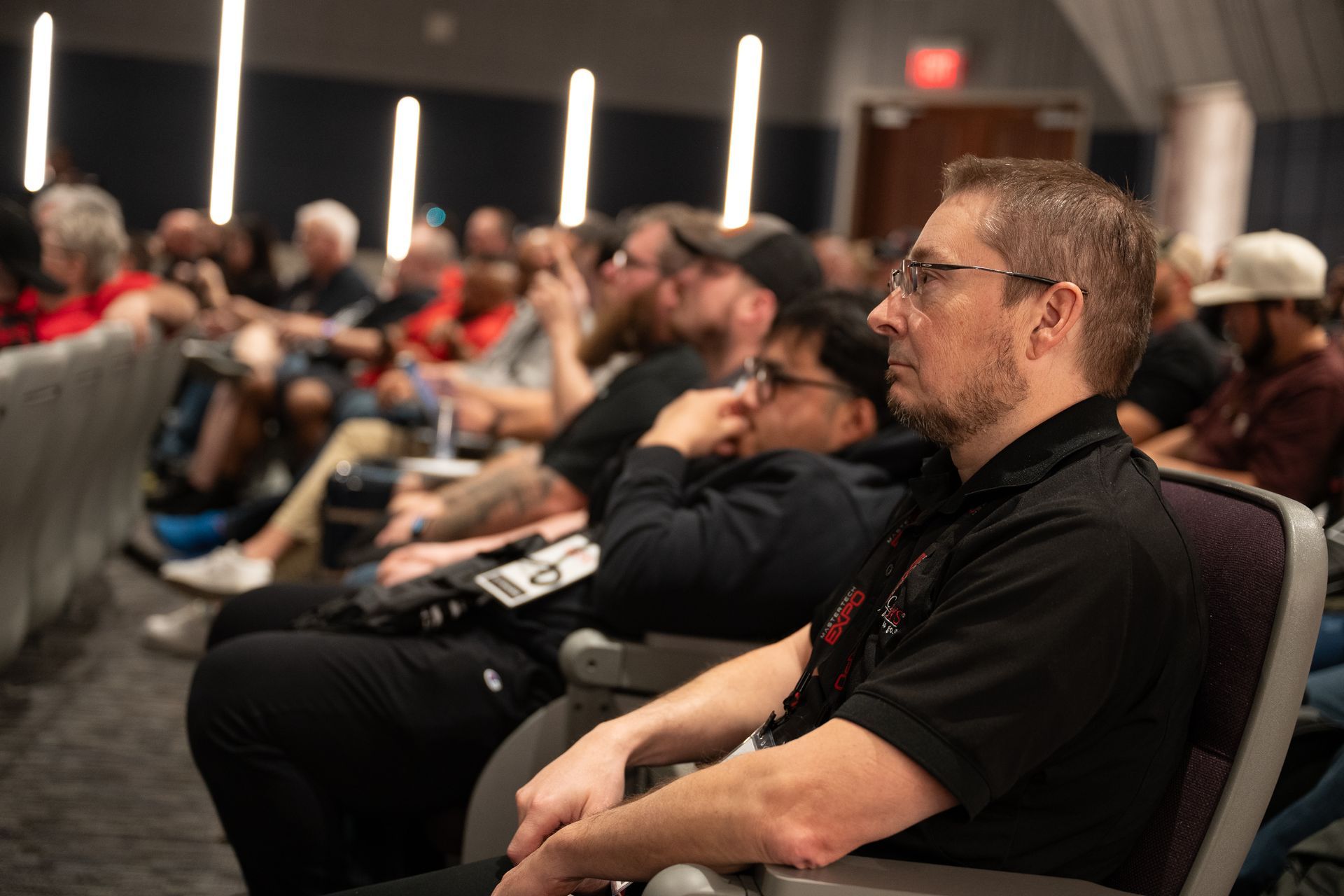 Audience members seated at a conference, listening to a presentation. A man in black shirt looks on.