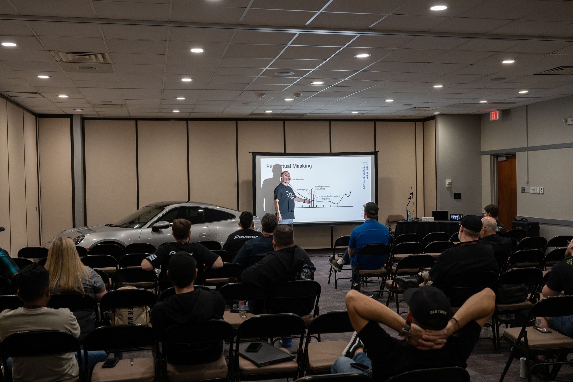 A man lectures in a classroom with a car displayed. Students sit, watching a presentation on a screen.