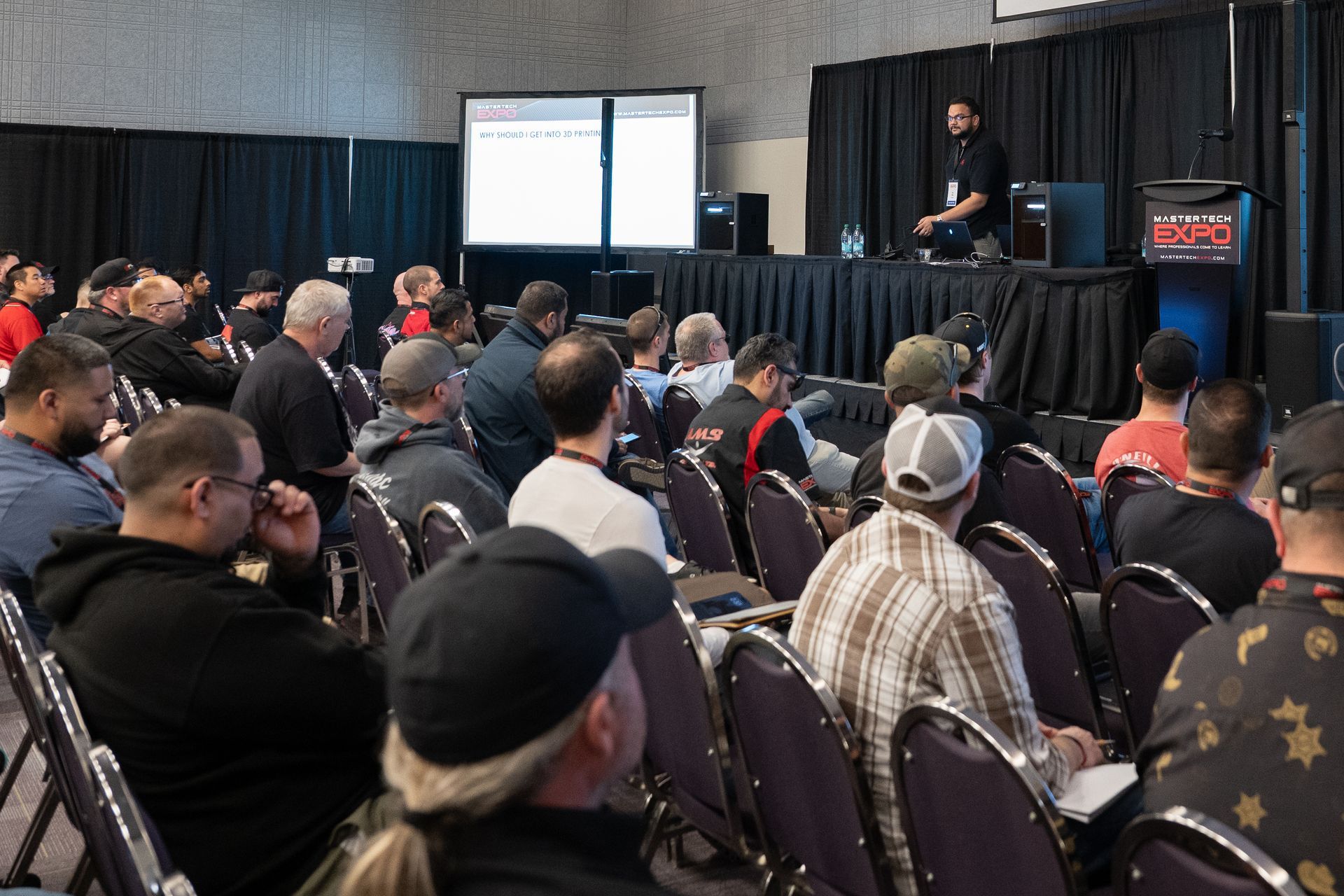 Audience watches a presentation at an event. A man speaks at a podium; screen displays a presentation.